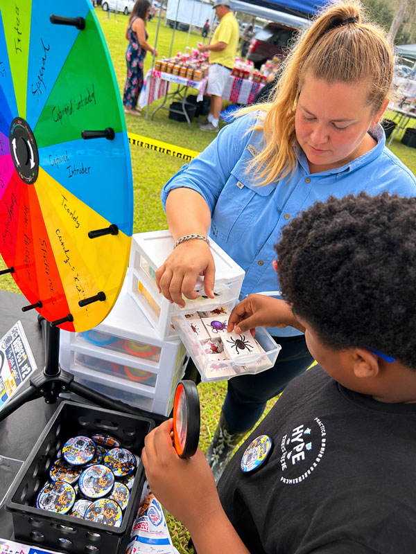 Bug Cops team member helping a child select a prize beside the colorful Junior Bug Cop prize wheel at the Florahome Fall Heritage Festival.