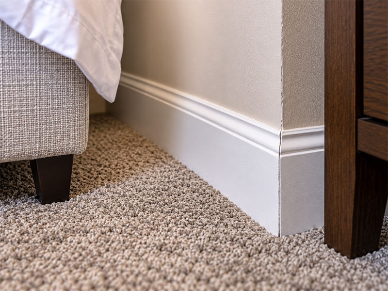 Close-up of a white bedroom baseboard and wall corner beside carpet and furniture, showing an area Bug Cops examines during bed bug control services.