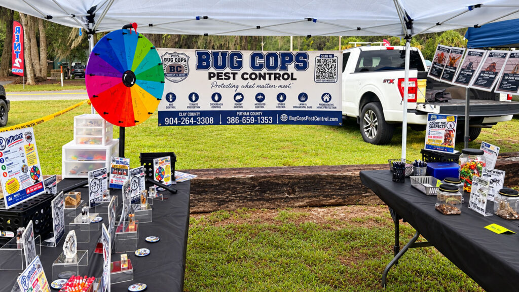 Bug Cops Pest Control booth at the Florahome Fall Heritage Festival in North Florida, featuring pest identification displays, a colorful prize wheel, and the company banner.
